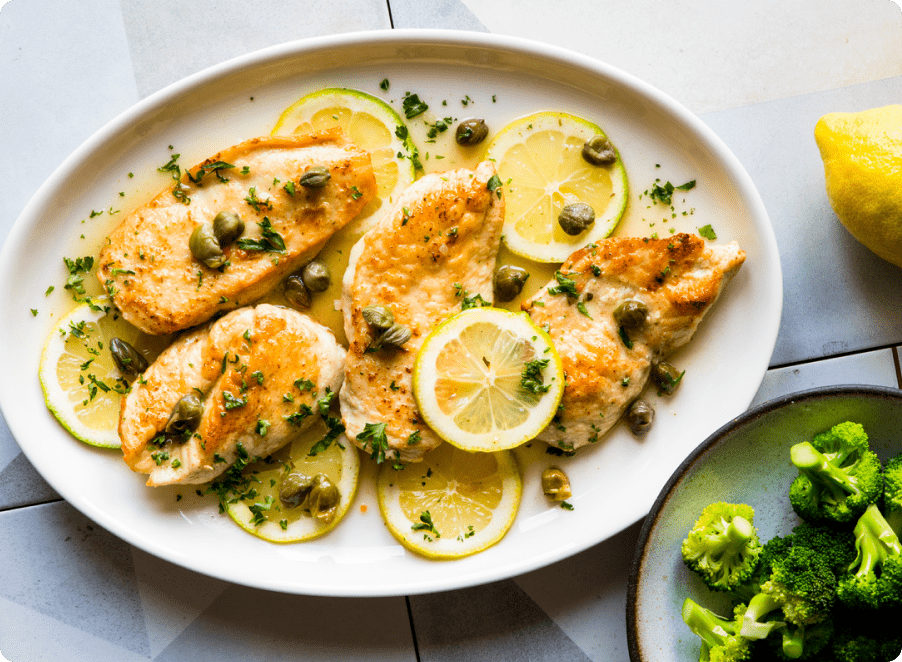 A plate of cooked juicy chicken breasts topped with lemon slices, capers and herbs